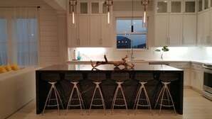 A modern kitchen with white cabinets, a central island with bar stools, pendant lighting, and a view of the outside at dusk.