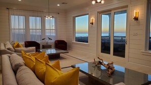 A modern living room with a sofa, chairs, a coffee table, and large windows offering a view of the sea.