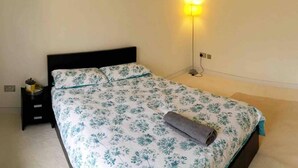 A bed with a floral bedspread, a bedside lamp, and a small table with a plant. - Hillcroft (King's Lynn)