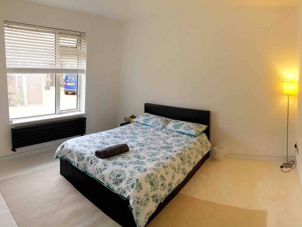 Une chambre à coucher avec un lit, une fenêtre avec des stores, un radiateur, une table de chevet et une lampe de sol.