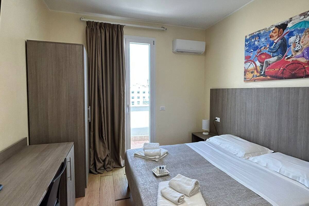 Una habitación de hotel con cama, escritorio, armario, ventana con cortinas, equipo de aire acondicionado y un cuadro en la pared.