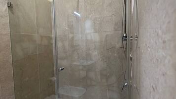 Cabina de ducha con paredes de vidrio, piso blanco, puerta de vidrio y ducha en la pared.