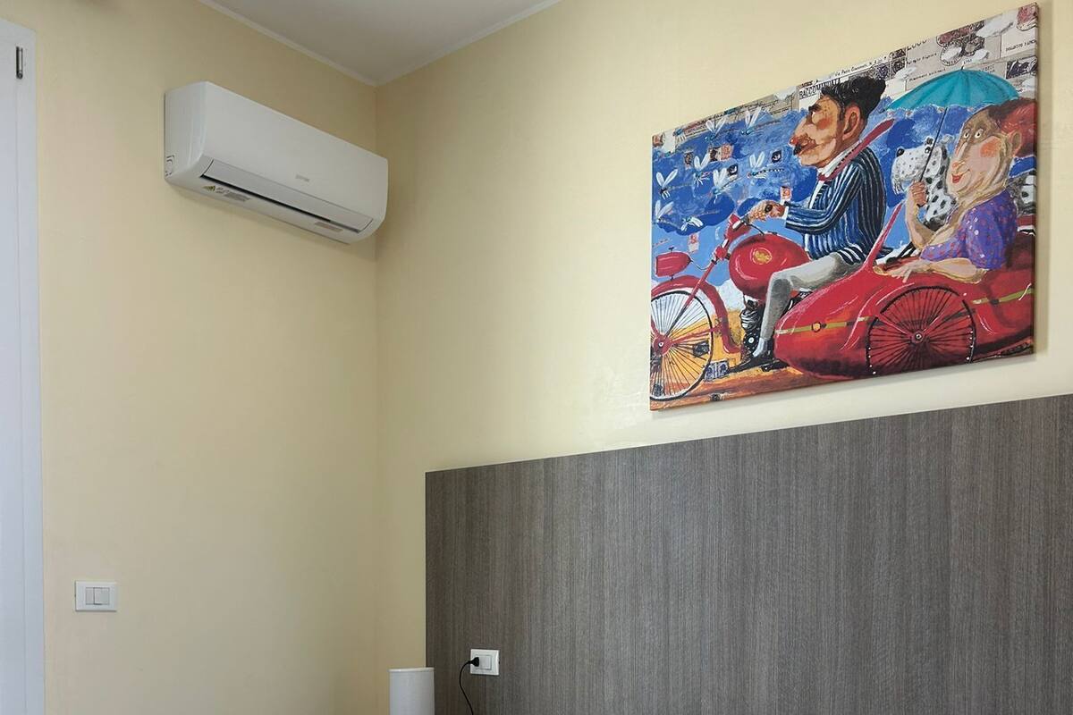 Una habitación de hotel con una cama, un cuadro en la pared, un aire acondicionado y una mesita de noche con lámpara.