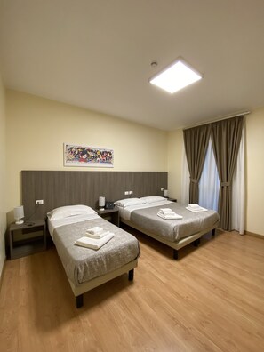 Une chambre d’hôtel avec deux lits simples, une tête de lit, une table de chevet avec une lampe, un tableau au mur et une fenêtre avec des rideaux.