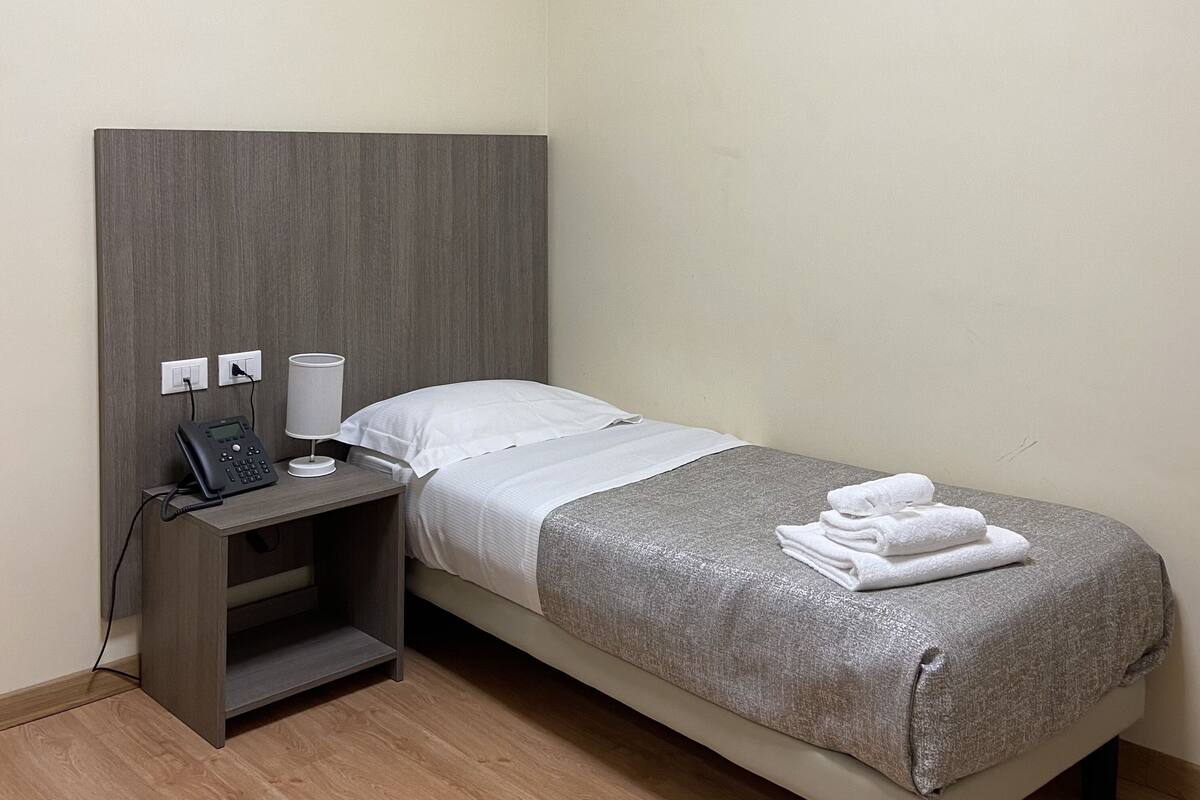 Una cama individual con cabecera gris, una mesita de noche con teléfono y lámpara, y un aire acondicionado instalado en la pared.