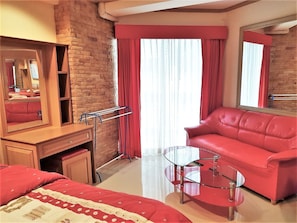 Quarto de hotel com parede de tijolos, sofá de couro vermelho, mesa de centro de vidro, escrivaninha de madeira e um espelho grande.