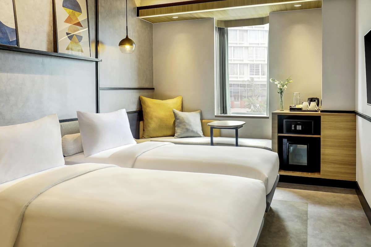 A hotel room with two beds, a small table, a microwave, and a window with a view of buildings.