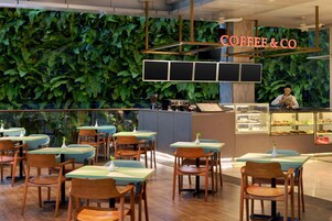 A modern coffee shop interior with green foliage walls, wooden tables, and a bar area.