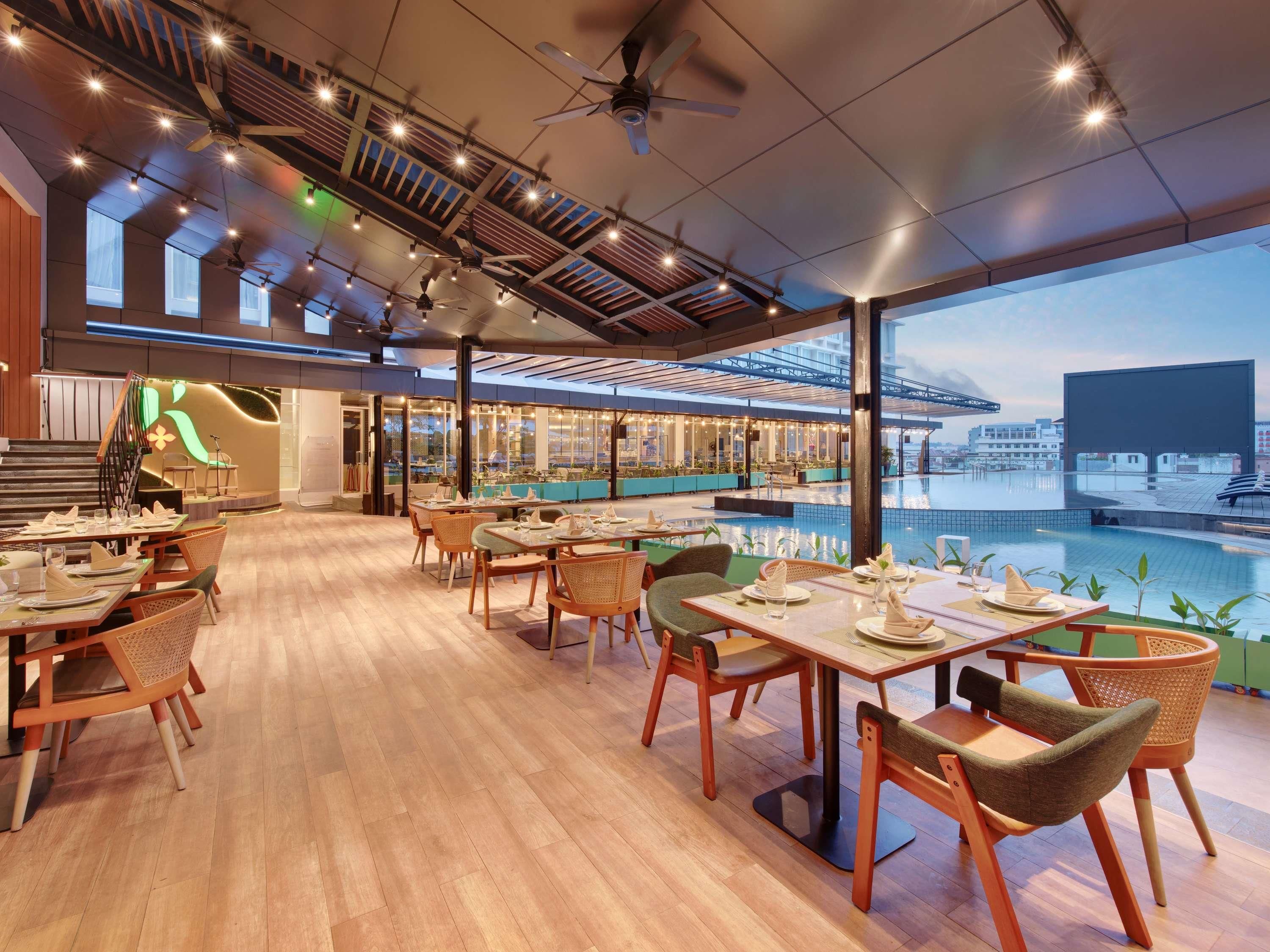 A spacious restaurant with wooden flooring, multiple dining tables, and chairs. There is a pool area with a view of the cityscape.