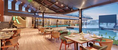 A spacious restaurant with wooden flooring, multiple dining tables, and chairs. There is a pool area with a view of the cityscape.