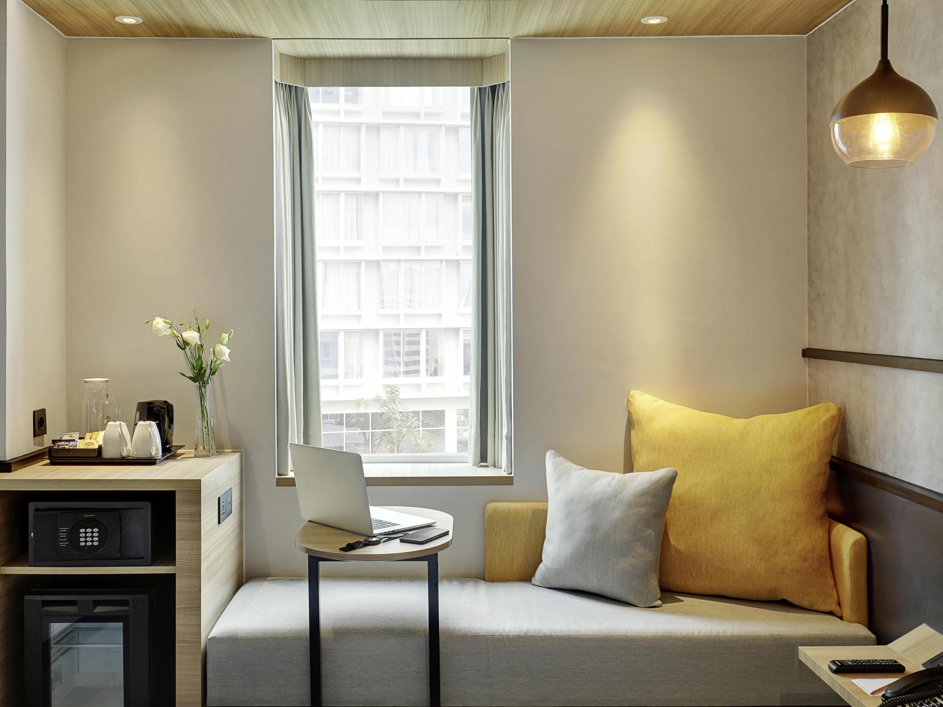 Una habitación de hotel moderna con sofá, laptop, cafetera y vistas de la ciudad a través de un amplio ventanal.