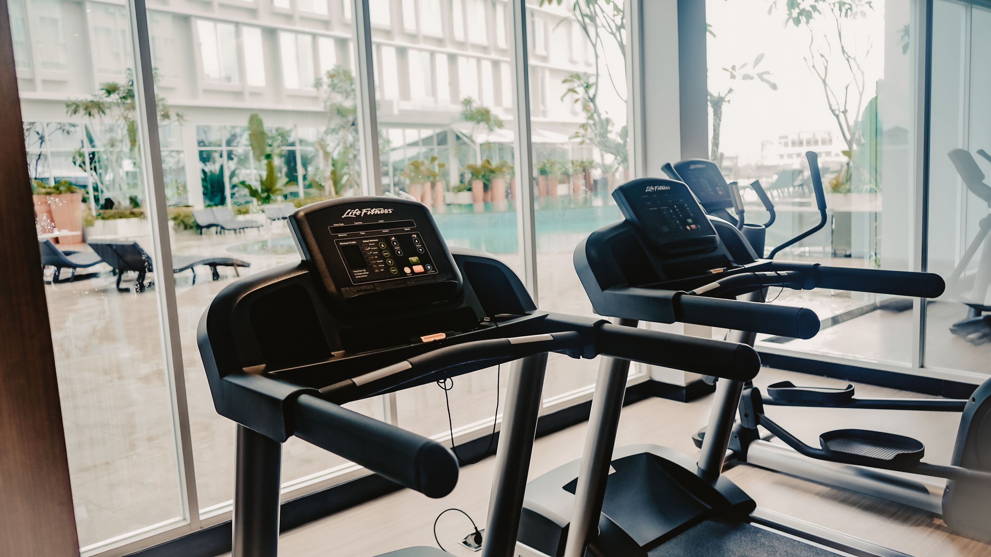 Una palestra con tapis roulant e attrezzature per esercizi, ampie finestre che offrono vista su un'area relax esterna e piante.