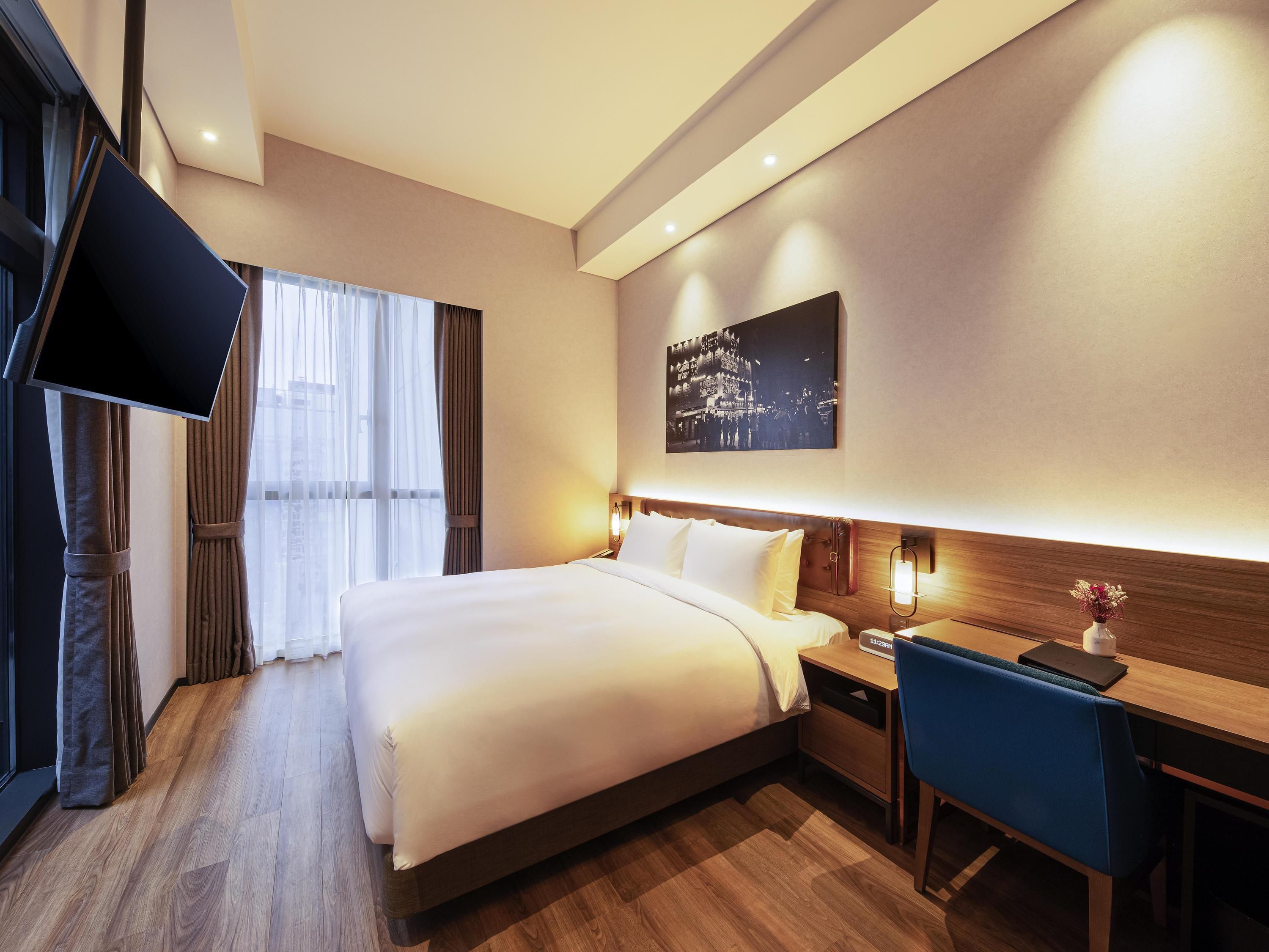 A hotel room with a bed, bedside table, desk, chair, TV, and a wall-mounted picture.