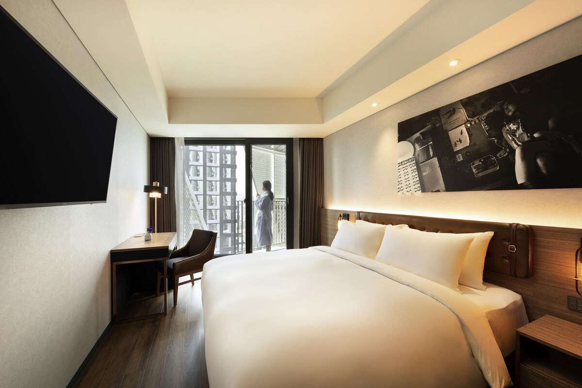Habitación de hotel con una cama grande, un escritorio y una silla. Se observa una ventana amplia con vistas a un edificio.
