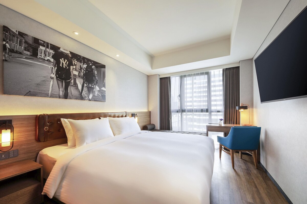 Una habitación de hotel moderna con una cama grande, un escritorio, una silla, un televisor de pantalla plana y un ventanal amplio.