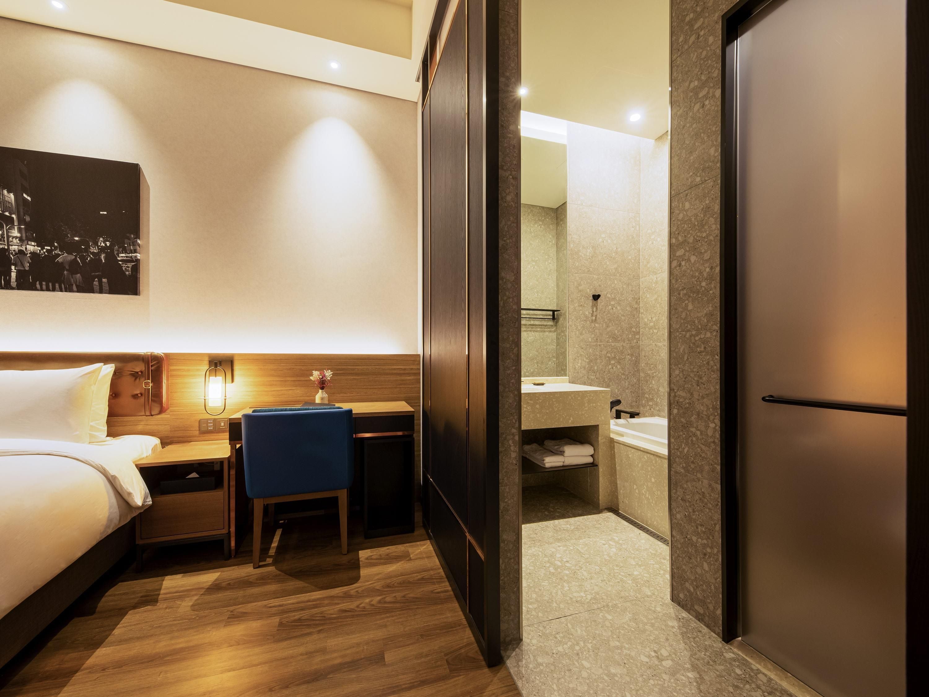 A hotel room with a bed, bedside table, chair, desk, and a bathroom with a sink and mirror.