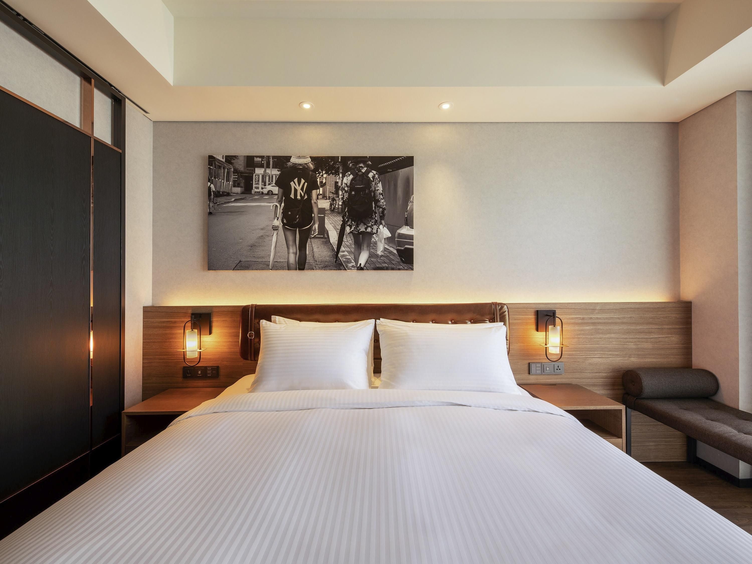 A modern hotel room with a large bed, two bedside tables with lamps, a bench, and a cityscape artwork on the wall.