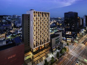 Egy modern hotel, melynek jellegzetes, geometrikus homlokzata van, és amelyet éjszaka egy forgalmas városi utca szegélyez.