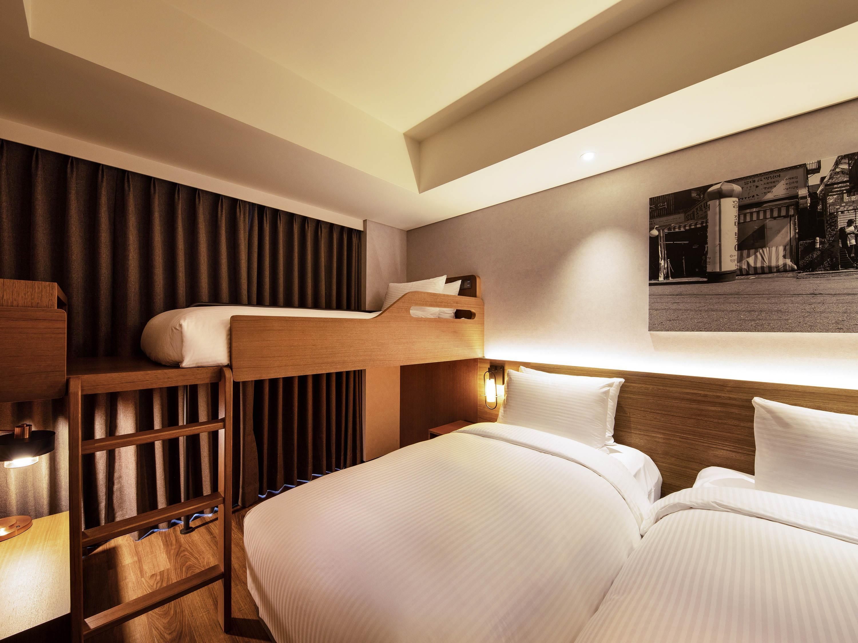 A hotel room with a bunk bed, a desk with a lamp, a bedside table, and a wall-mounted light fixture.