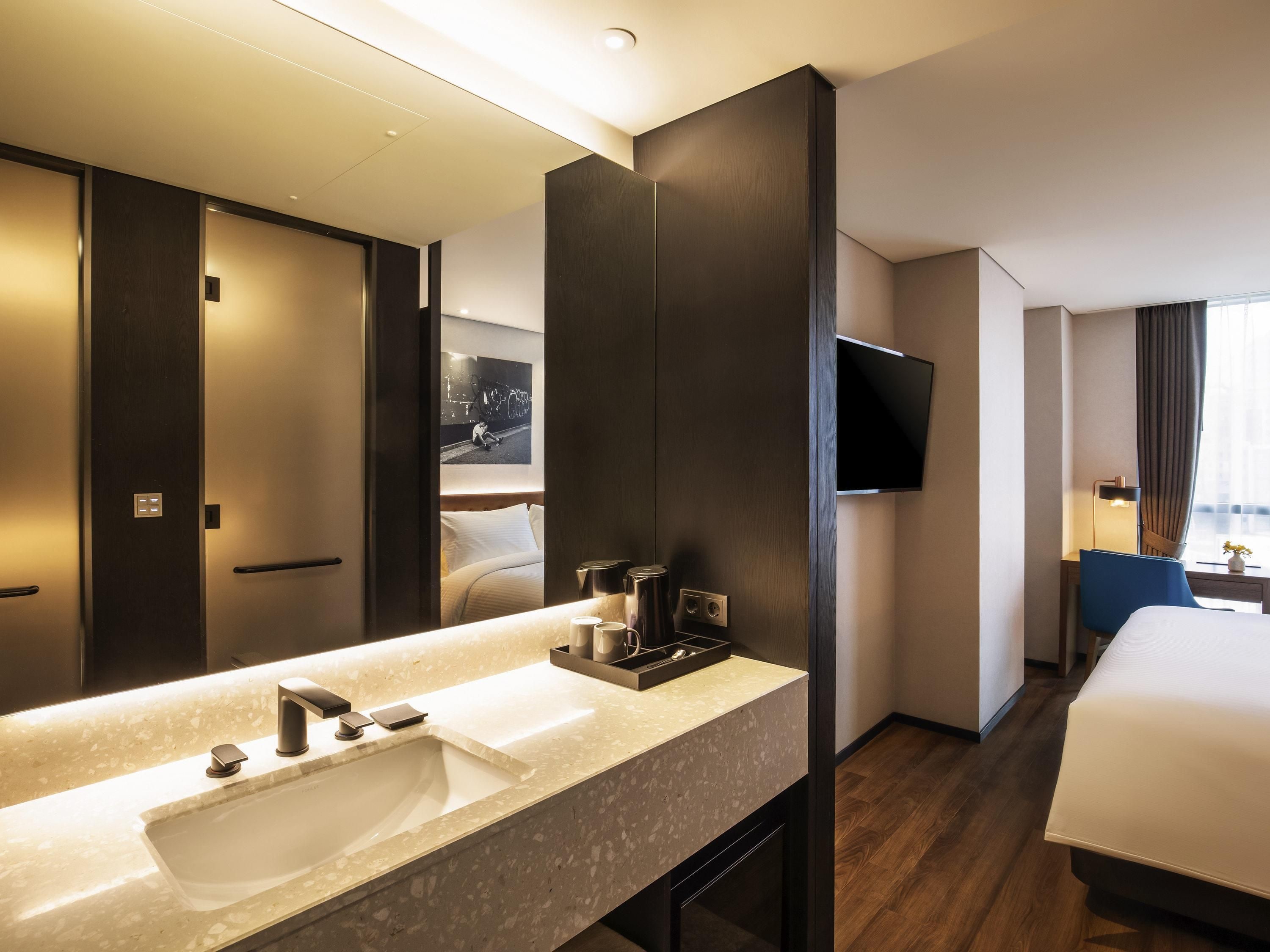 A modern hotel room with a large bed, a bathroom with a double sink, a flat-screen TV, and a large mirror.