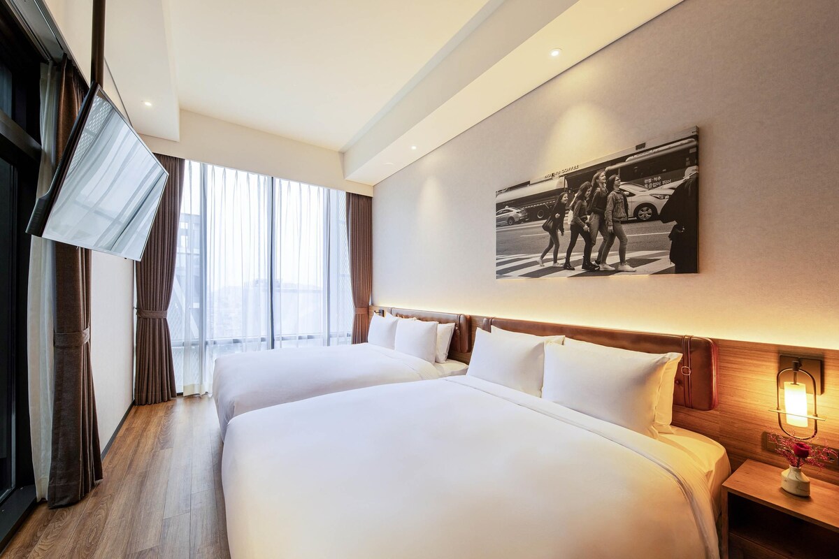 Habitación de hotel con dos camas, un ventanal con cortinas, un cuadro en la pared y una mesita de noche con lámpara.