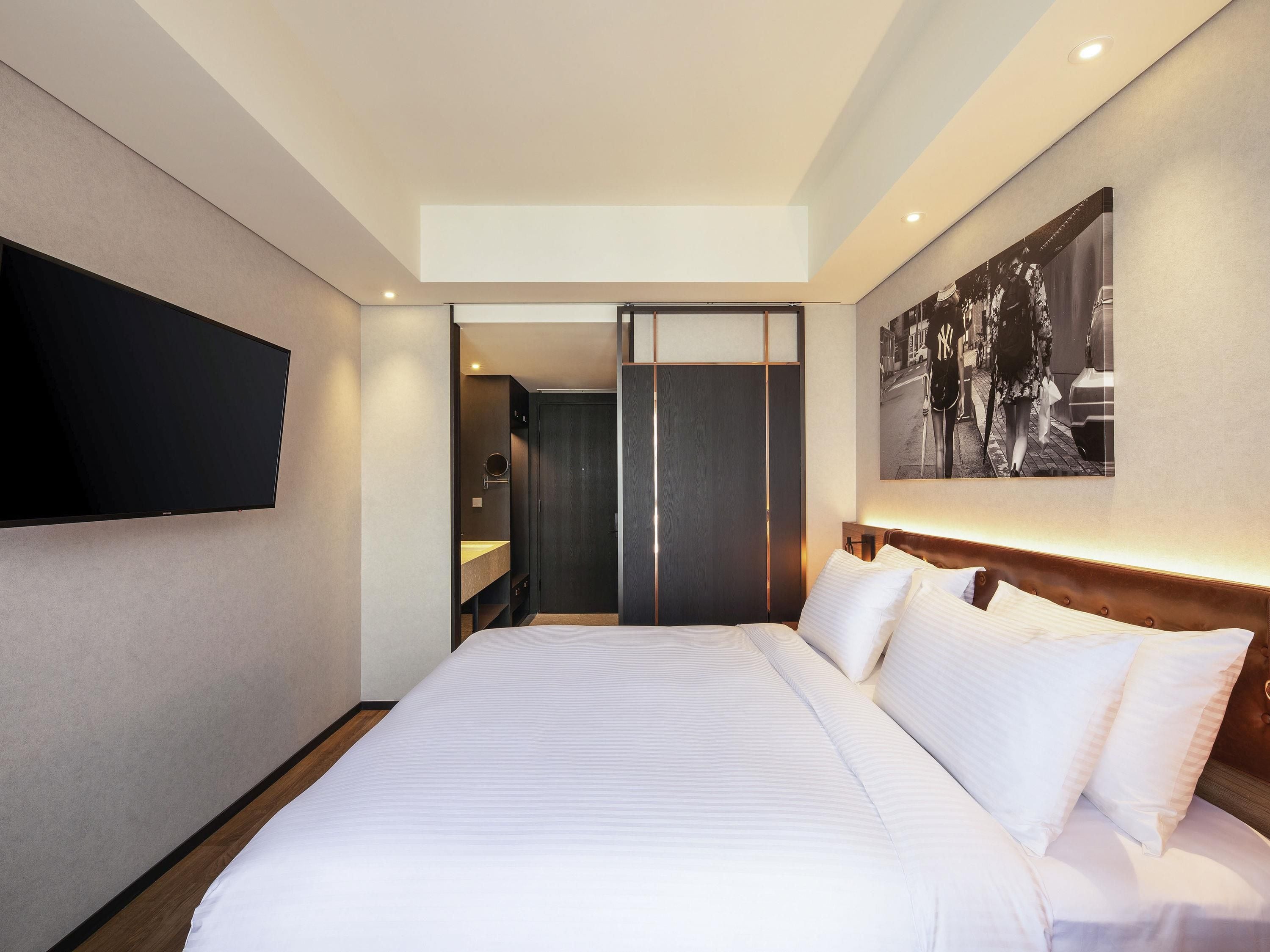 Una habitación de hotel con una cama grande, un televisor de pantalla plana montado en la pared, un cabecero de madera y un cuadro enmarcado.