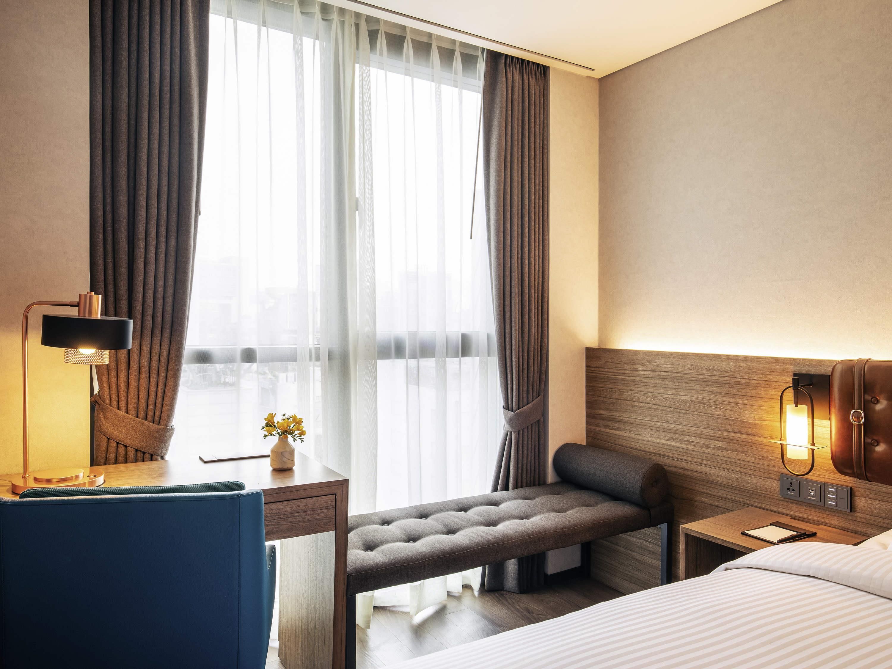 A hotel room with a bed, bedside table, lamp, bench, and a view through sheer curtains.
