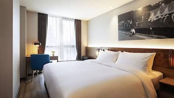 Une chambre d’hôtel avec un grand lit, un bureau, une chaise, une table de chevet, une lampe et une grande fenêtre avec des rideaux.