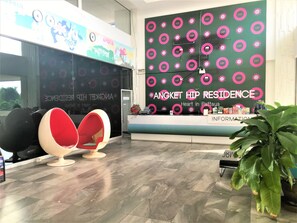 Ein Empfangsbereich mit einer großen Wand, auf der der Text 'Angket Hip Residence Heart in Pattaya' steht. Im Vordergrund stehen zwei rote Stühle, eine Rezeption mit Informationsbroschüren und eine Zimmerpflanze auf der rechten Seite.