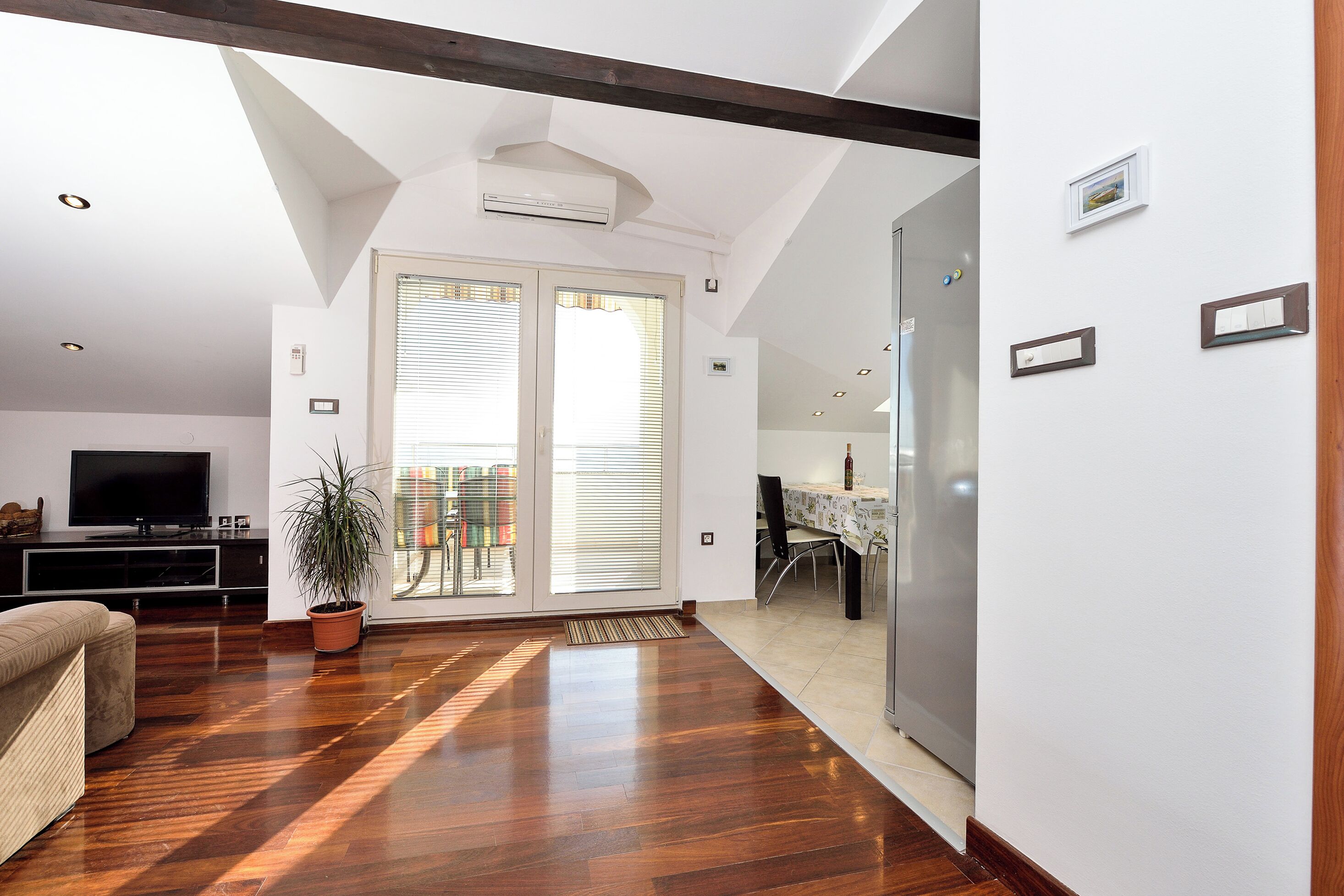 A spacious living area with hardwood flooring, a sofa, a television, and a potted plant.