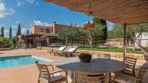 Un espace aménagé au bord de la piscine, avec une table ronde et des chaises, un luminaire suspendu et une vue sur une maison dotée d’un mur en pierre et de verdure.
