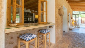 Un intérieur rustique avec un comptoir de bar en bois, deux tabourets dotés de coussins à motifs et un sol en pierre.