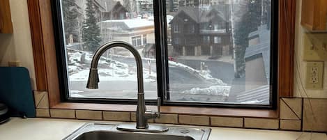 Une cuisine dotée d’un évier en acier inoxydable, de meubles en bois et d’une fenêtre donnant sur un paysage enneigé avec des bâtiments.