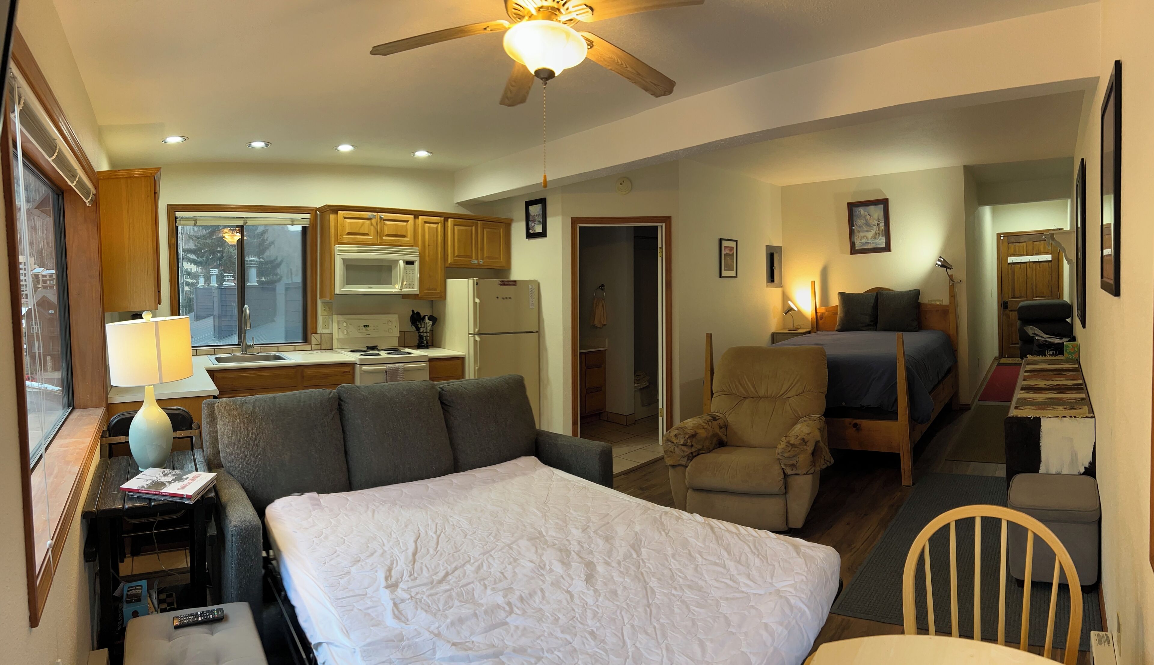 A bedroom with a large bed, a chair, a desk with a lamp, and a view of a kitchen and bathroom.