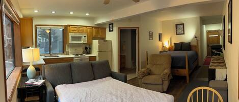 A bedroom with a large bed, a chair, a desk with a lamp, and a view of a kitchen and bathroom.