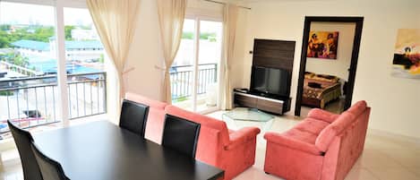 Een moderne woonkamer met een eettafel, roze banken, een televisie en een balkon met uitzicht op gebouwen en groen.