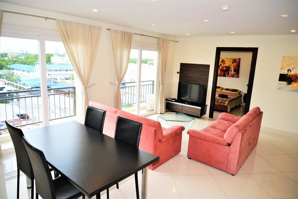 Una sala de estar moderna con una mesa de comedor, sofás rosados, un televisor y un balcón con vistas a edificios y follaje.