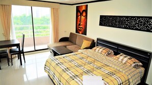 A hotel room with a large bed, a sofa, a dining table, and a view of greenery through the window. - Angket Condominium Corner Apartment With Large Balcony (Pattaya)