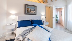 Une chambre avec un grand lit, des murs blancs et un plafond en bois. Un tableau encadré est accroché au mur, et il y a une table de chevet avec une lampe.