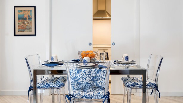 Un coin repas avec une porte blanche, un tableau encadré au mur, et une table agrémentée de chaises transparentes et de coussins à motifs bleus.