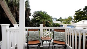 A balcony with two wooden chairs and a small round table, surrounded by white railings and palm trees.