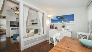 A cozy dining area with a wooden table and chairs, a window seat with cushions, and a painting of a beach scene on the wall.