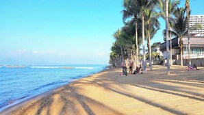 A beach with palm trees, a clear sky, and a building with a sign. - Sea View Studio Apartment Jomtien Beach Condominium A3 (Pattaya)