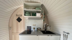 A compact kitchen with a built-in oven, a sink, and a small countertop. There is a wooden door with a towel hanging on a hook, and a shelf above the countertop holding dishes and utensils. - Iglu Camp Templin (Templin)