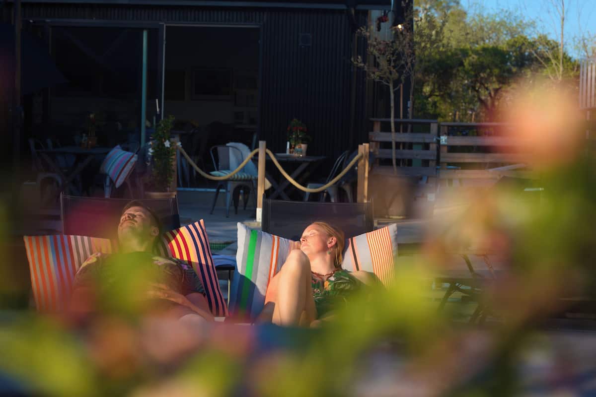 Two people reclining on lounge chairs with striped pillows, a wooden fence, and a building in the background.