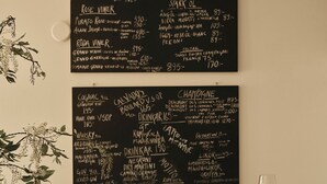 L’intérieur d’un restaurant présente un tableau noir affichant une carte des vins et des boissons, une table en bois avec des verres et des assiettes, et des chaises en métal noir.