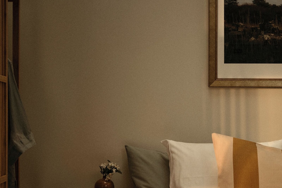 A neatly made bed with white and brown bedding, a small bedside table with a vase of flowers, and a framed picture on the wall.