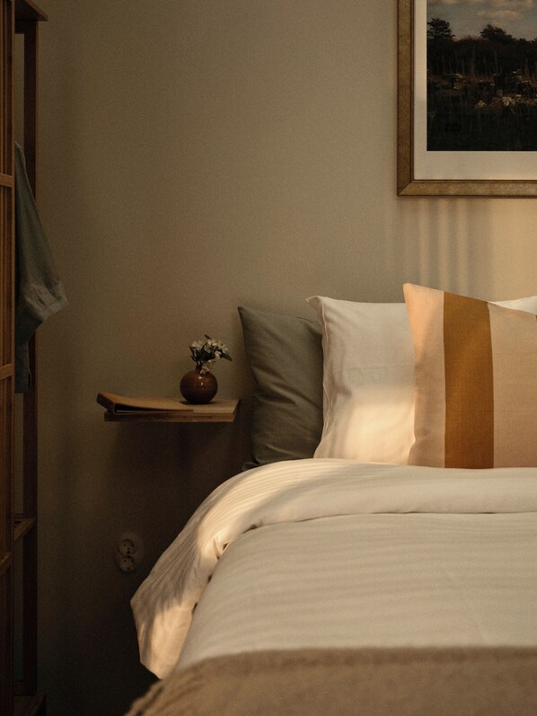A neatly made bed with white and brown bedding, a small bedside table with a vase of flowers, and a framed picture on the wall. - Hotell Persnäs (Löttorp)