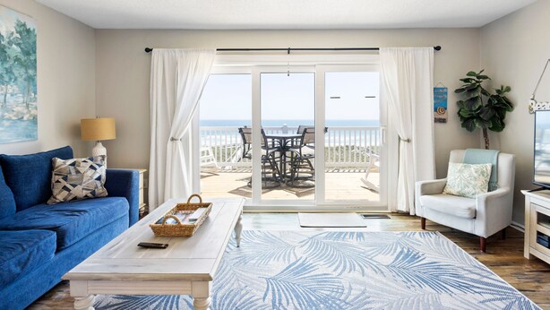 A room with a bed, a blue sofa, a white armchair, a table with a lamp, and a view of the sea through a sliding glass door.
