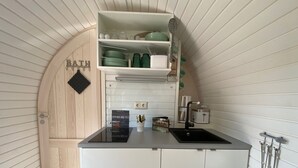 Cocina compacta con horno empotrado, lavamanos y una pequeña encimera. Se observa una puerta de madera con una toalla colgada en un perchero, y un estante sobre la encimera con vajilla y utensilios.